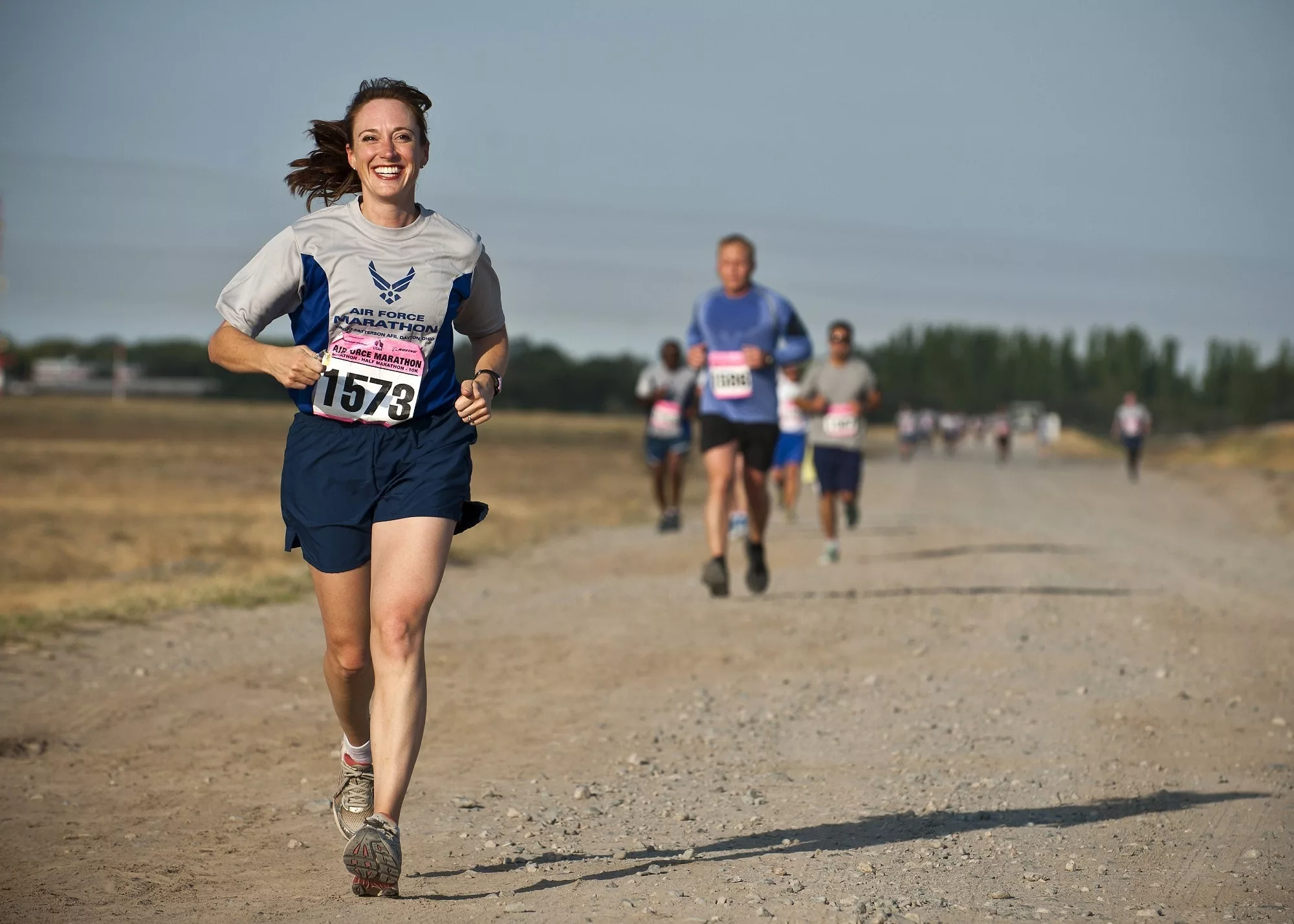 Woman running a marathon for a charity fundraiser. She's running on a dirt road, ten metres ahead of another runner She’s smiling with her ponytail whipping up into the air behind her head. This is a decorative image for a fundraising copywriter service page aimed at fundraising managers of nonprofits and charities.